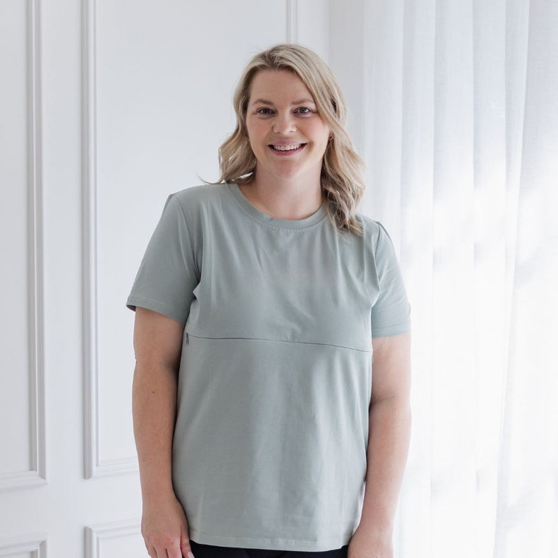 A smiling mum breastfeeds her baby girl easily in a light sage nursing t-shirt.