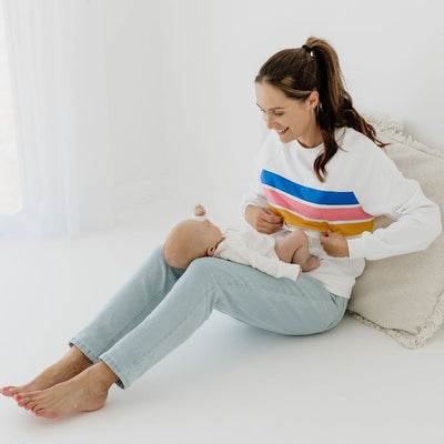 A young mum unzipping her nursing jumper as she smiles at her baby resting on her legs.