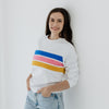 A young woman leaning against a wall as she models a white nursing sweater that has three colored stripes.