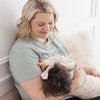 A smiling mum breastfeeds her baby girl easily in a light sage nursing t-shirt.