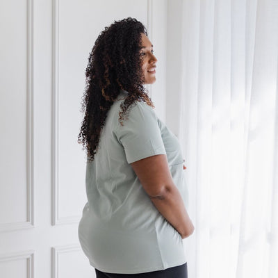 A pregnant woman holds her growing bump while modeling the fit of a breastfeeding t-shirt.